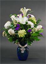 A memorial design with white lilies, snow white roses, with white carnations, purple statice, green hydrangeas and mixed green all arranged in a cobalt blue vase and tied with a keepsake angel medallion.